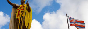 King Kamehameha Statue with lei and the Hawaiian Flag with blue sky and white clouds.