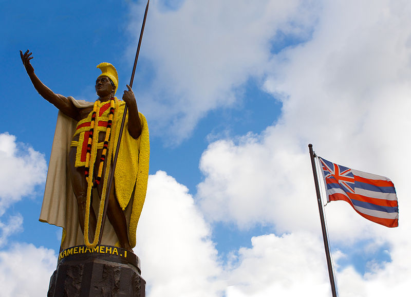 King Kamehameha Statue with lei and the Hawaiian Flag with blue sky and white clouds.